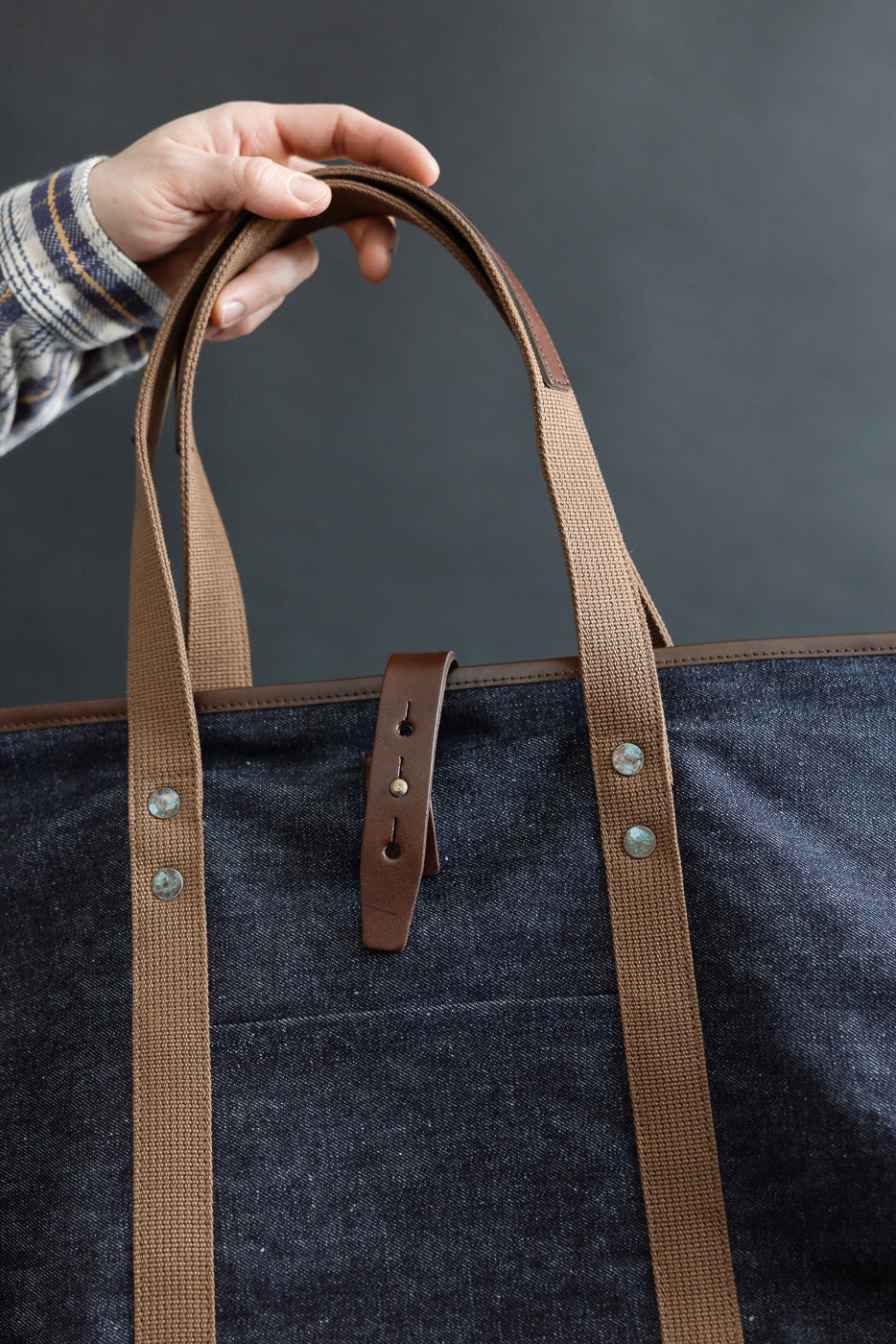 Close-up of front side of indigo denim tote bag with brown canvas handles, leather strap, and metal rivets held by male model.