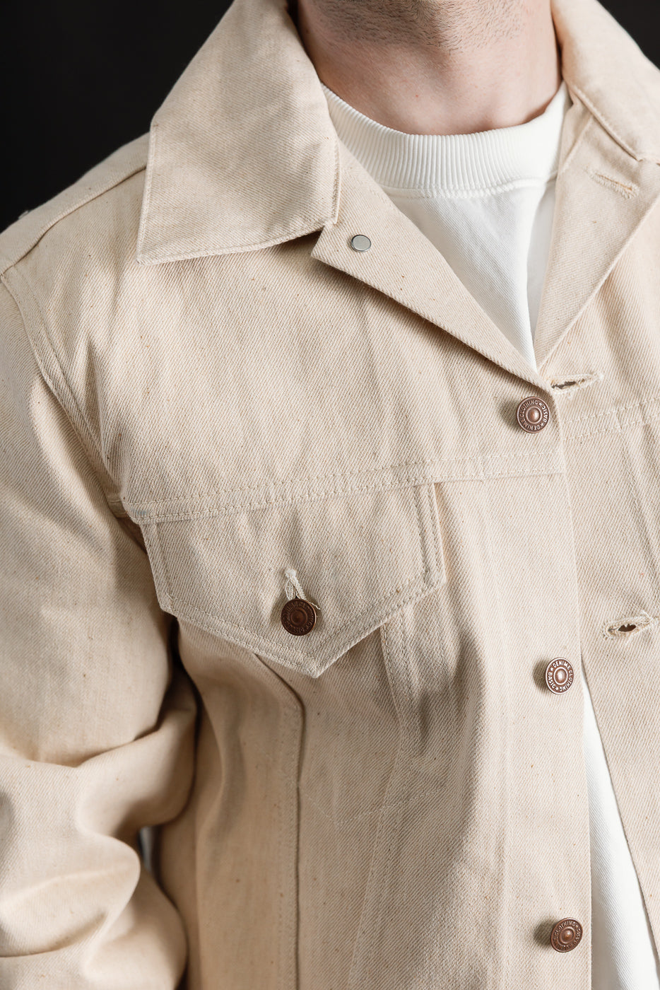 Close-up front detail of ecru selvedge denim jacket on male model, showing collar, pocket, and buttons; model is 5'9