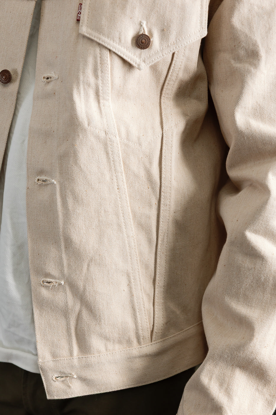 Close-up of male model wearing ecru denim jacket, front torso view, showing chest pocket, buttons, and fabric texture. Model is 5'9