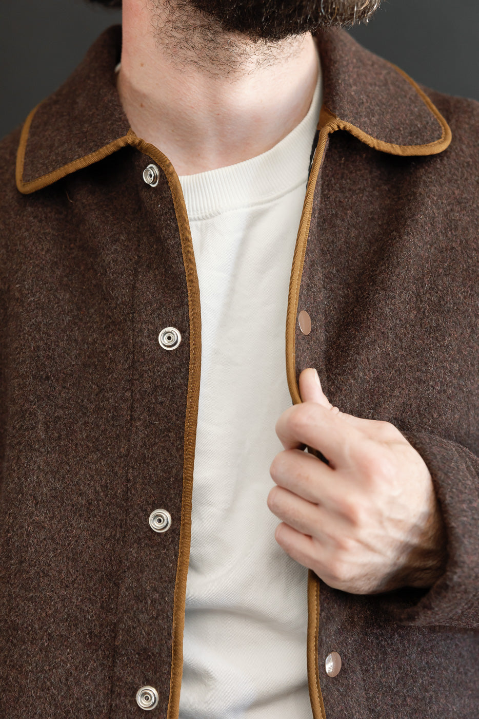 Close-up front view of a brown wool jacket with tan piping and silver snap buttons on a male model.