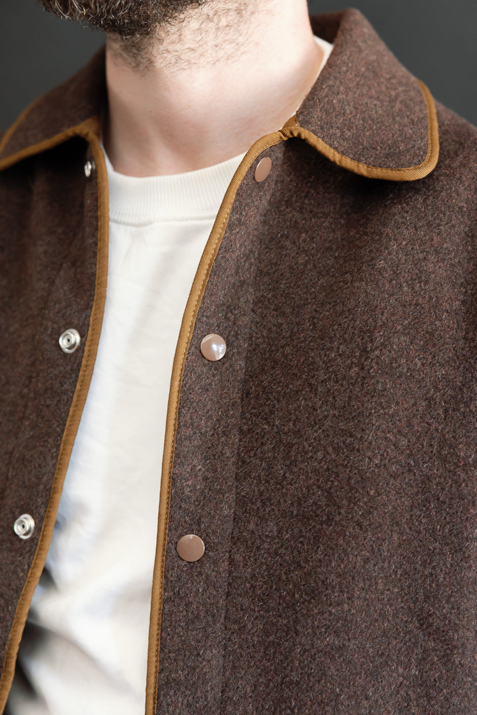Close-up front view of brown wool jacket with tan piping and metal snap buttons on a male model.