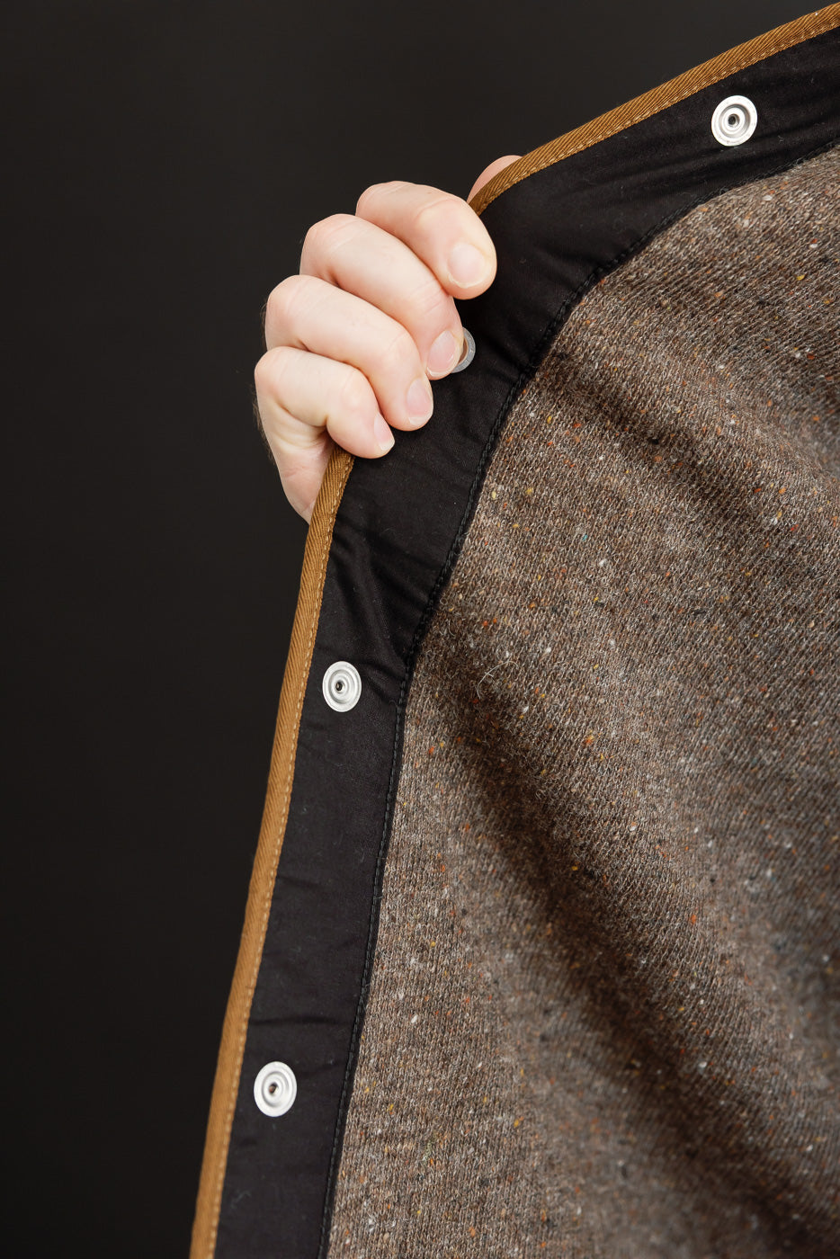 Close-up of the brown textured fabric and black interior lining of a garment edge with silver snaps.