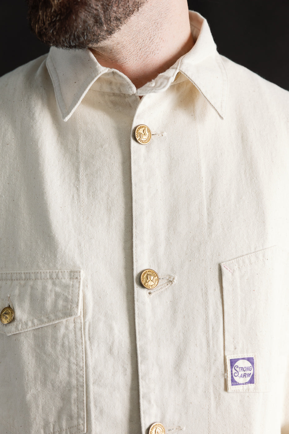 Close-up of front chest of ecru house painter's jacket, gold buttons, pointed collar, Strong Arm patch, on male model 5'9