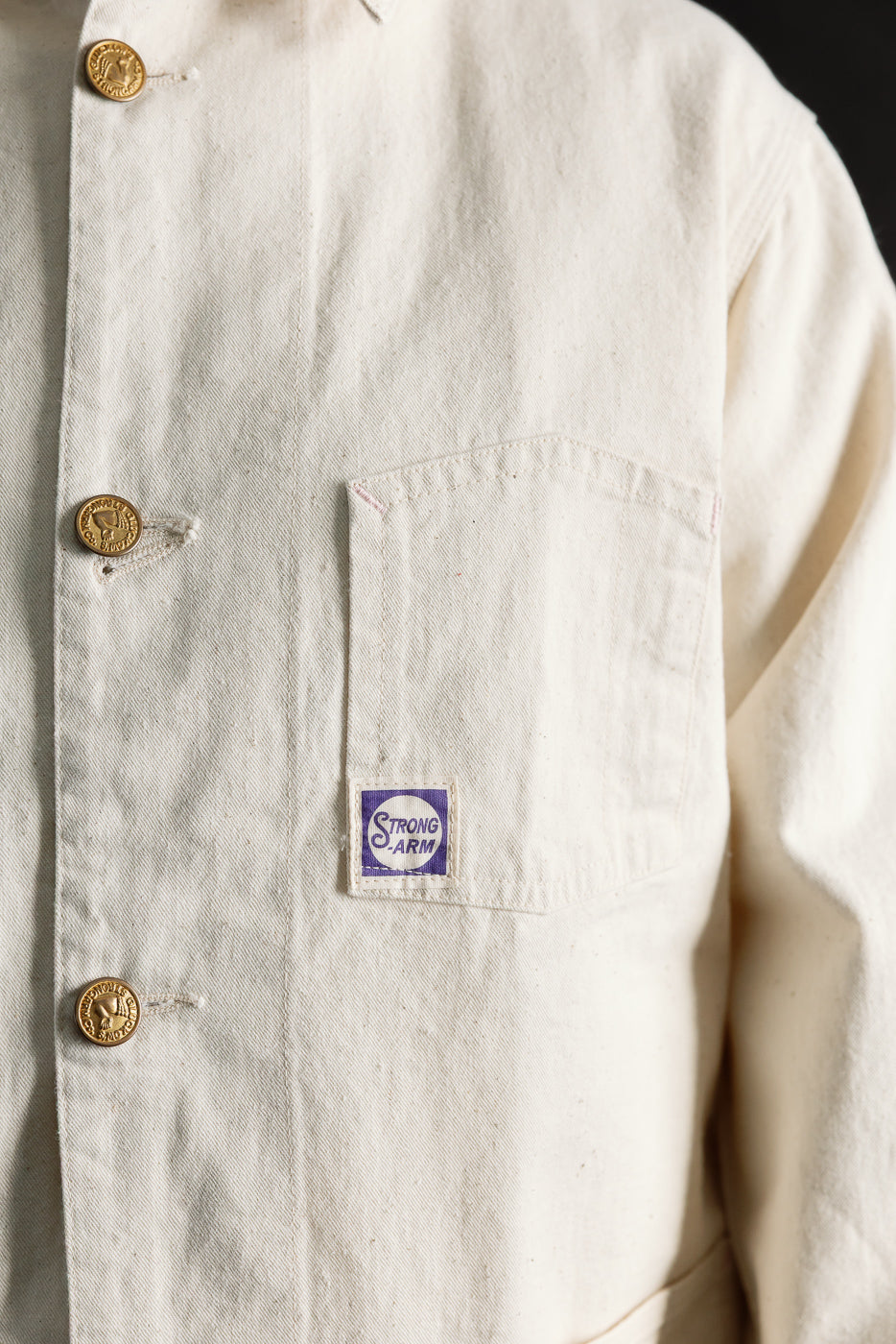 Close-up of the front chest pocket and buttons on an ecru men's painter jacket; visible 'Strong Arm' tag, textured fabric, and brass button details.