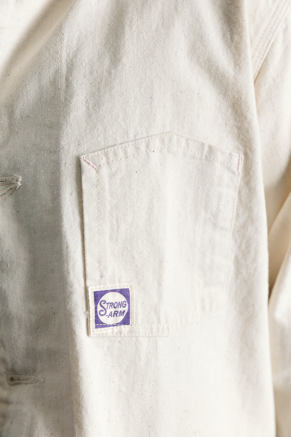 Close-up of front left chest pocket on ecru painters jacket, Strong Arm tag, fabric texture visible