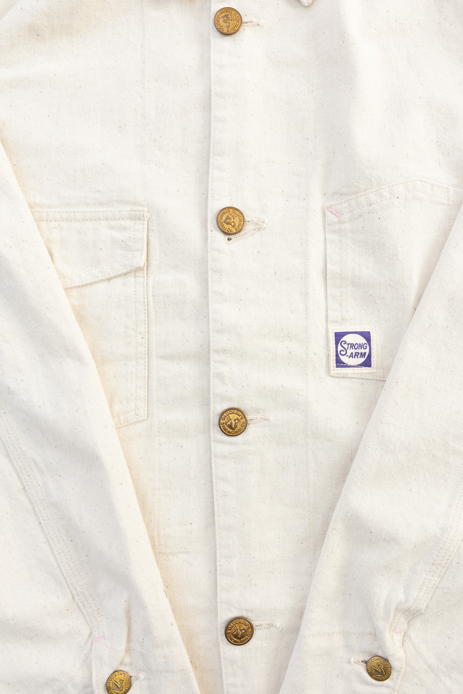 Close-up of front of ecru painter's jacket showing brass buttons, patch pockets, and Strong Arm tag