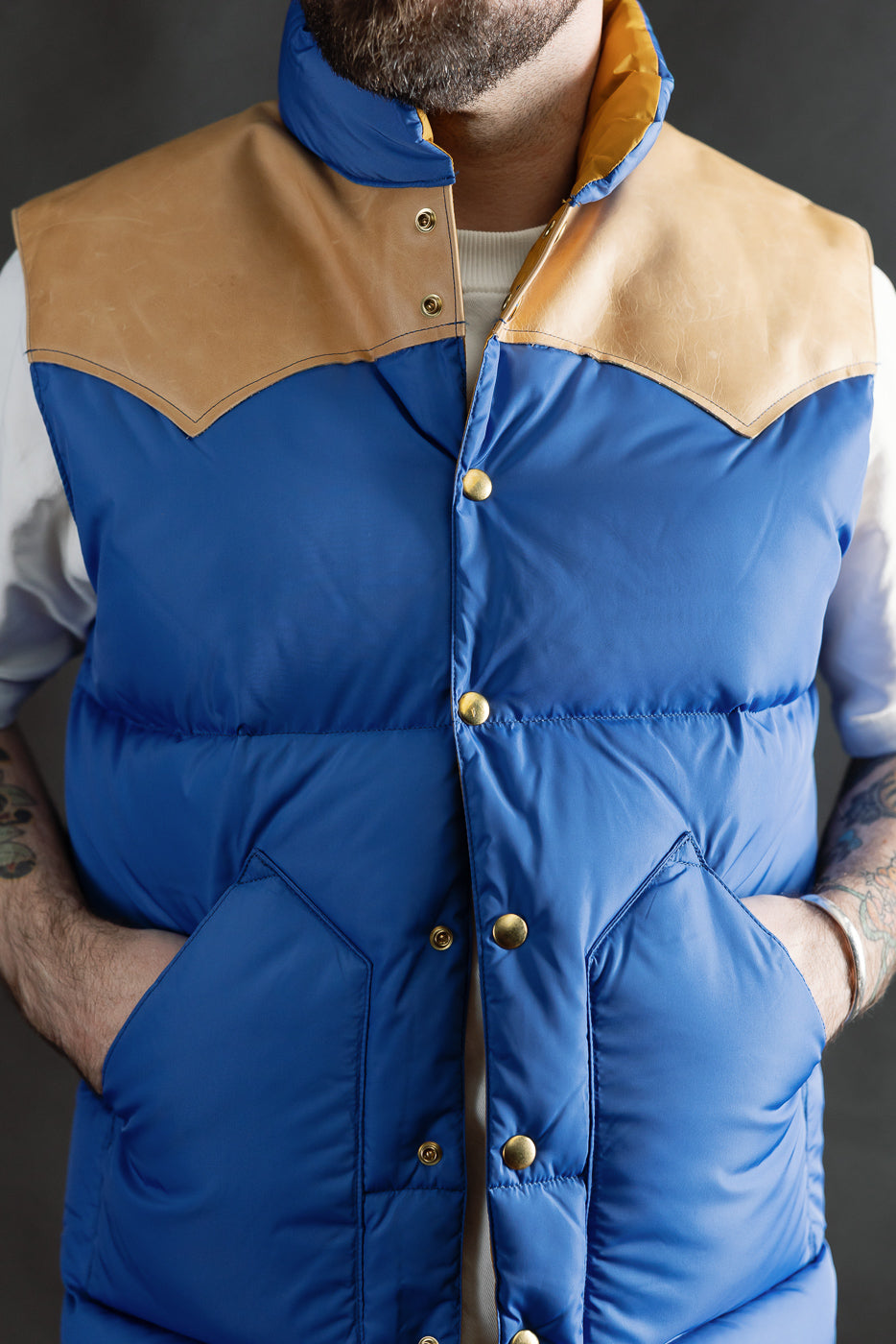 Close-up front view of blue nylon down vest with tan yoke, brass snaps, modeled by male 5'9