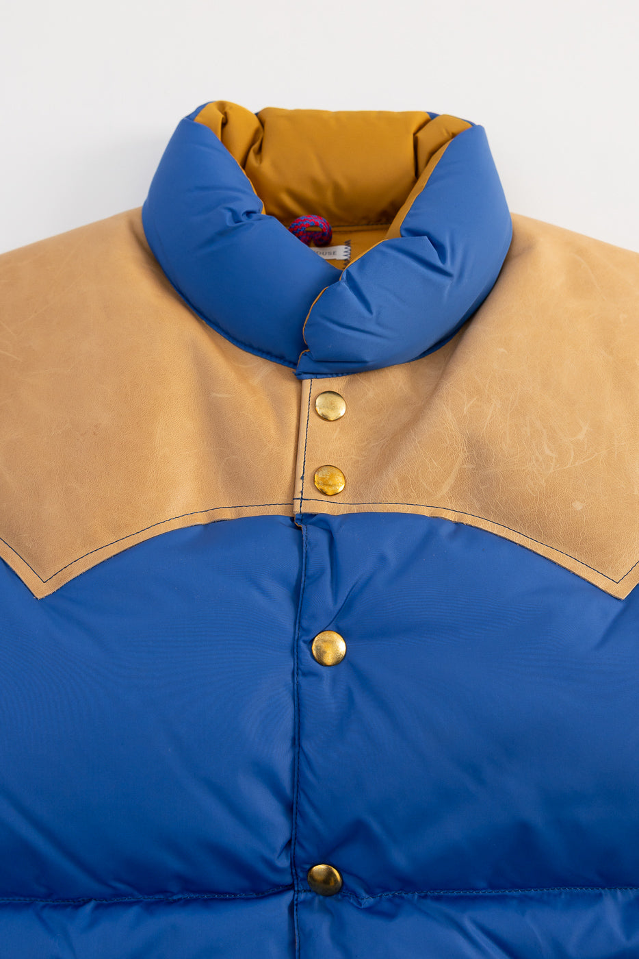 Close-up front of blue nylon down vest, tan yoke, gold snap buttons, Dant contemporary casual apparel