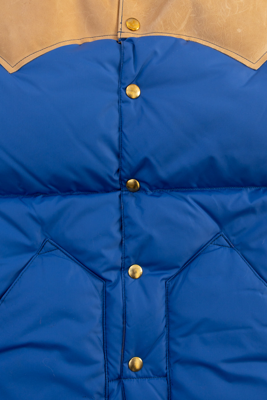 Close-up front detail of blue nylon down vest with brass snap buttons and tan yoke