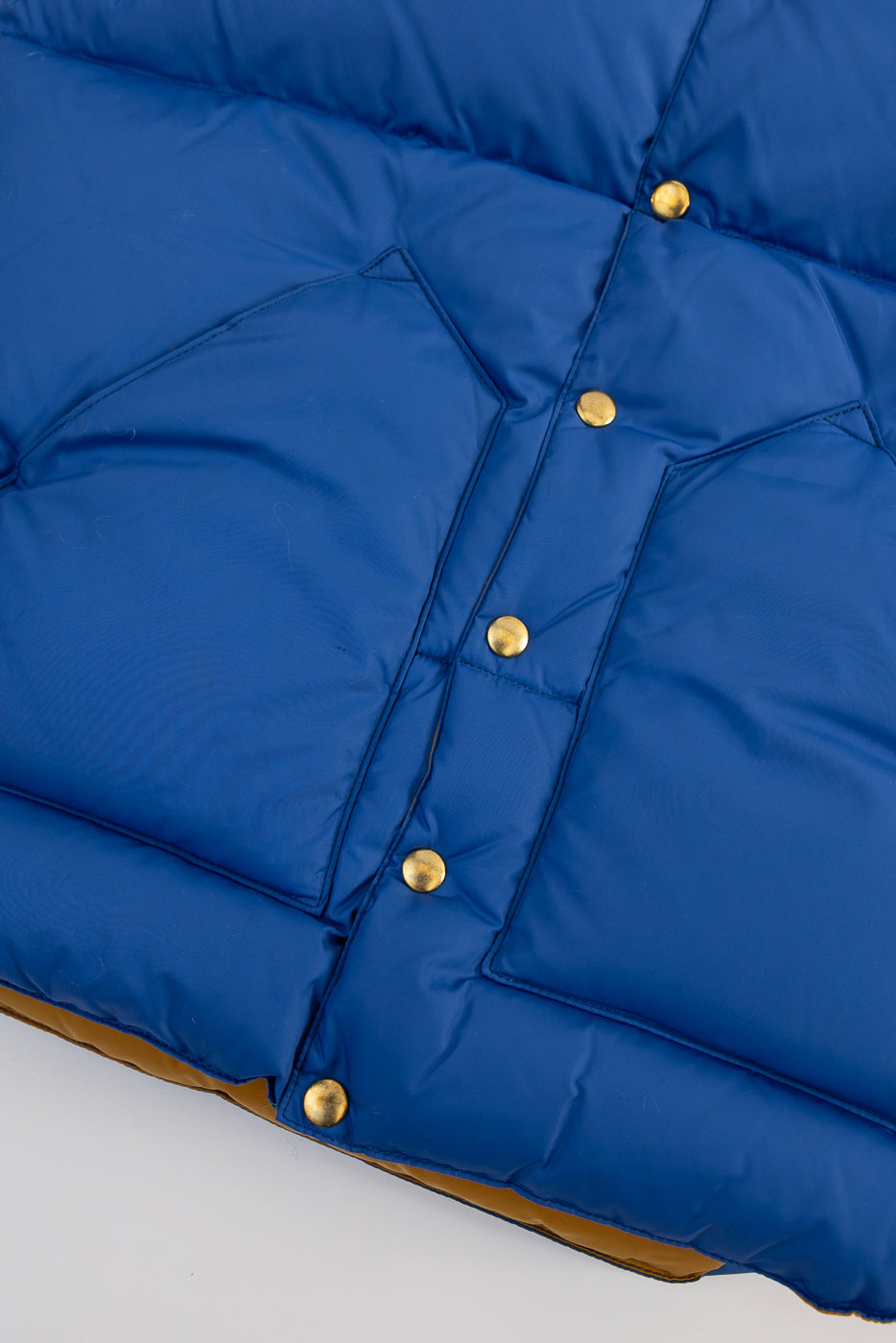 Close-up of front lower section of blue nylon down vest showing gold snap buttons and pocket details