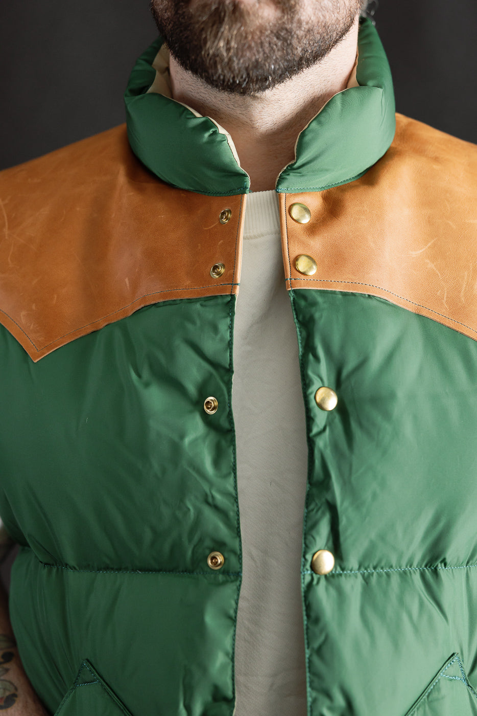 Close-up front of bottle green nylon down vest with tan leather yoke, gold snaps, on male model 5'9