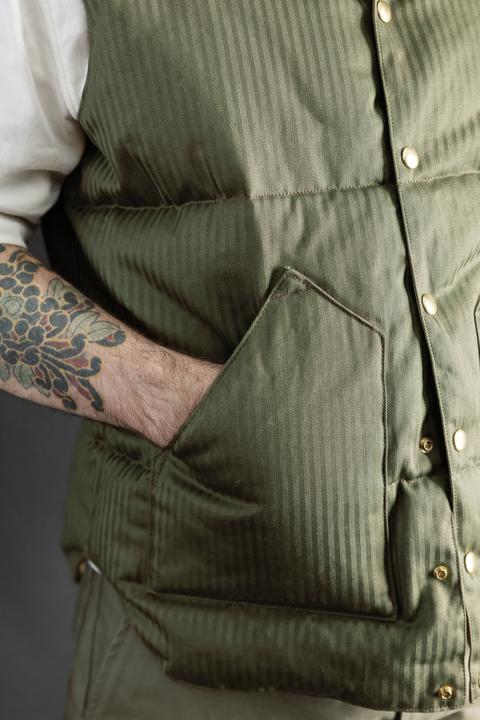 Close-up of front left side of OD green down vest on male model, showing snap buttons, pocket, and herringbone texture; model is 5'9