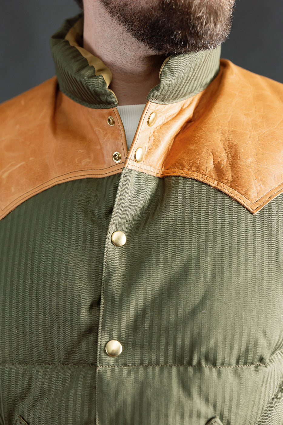 Close-up front of Rocky Mountain Warehouse OD green down vest on male model, showing collar, snap buttons, and tan yoke. Model is 5'9