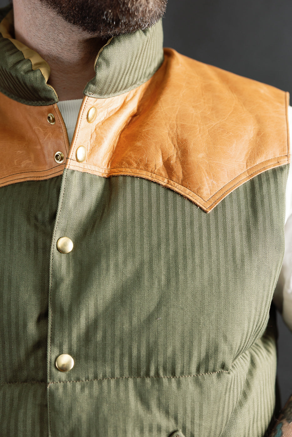 Close-up of front chest and collar on OD green down vest, tan yoke, brass snaps; on male model, 5'9