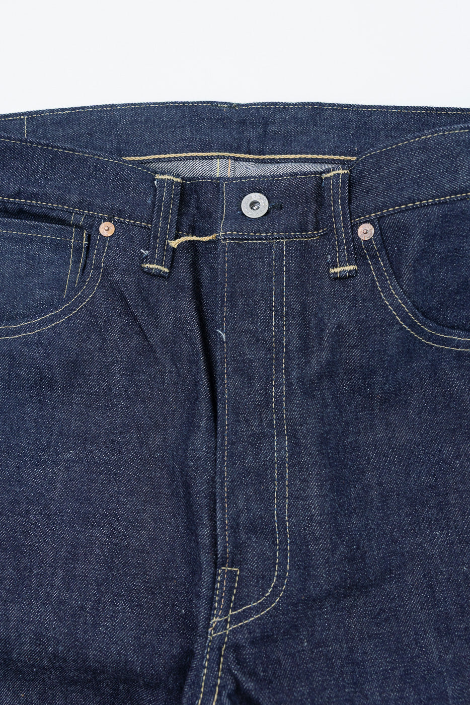 Close-up front view of dark indigo selvedge denim jeans showing waistband, button, and fly.