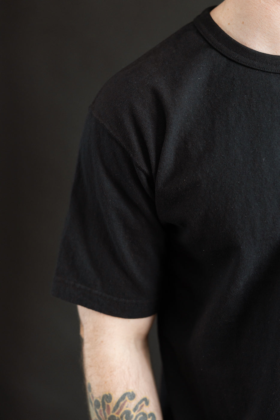 ELS Recycled Cotton Slub Crewneck T-Shirt - Black: Close-up of an upper torso wearing a plain black T-shirt, showcasing its modern athletic fit and textured fabric with a colorful tattoo in view.