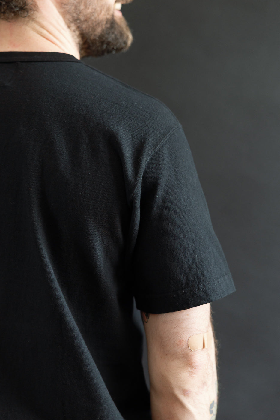 Man wearing the ELS Recycled Cotton Slub Crewneck T-Shirt - Black, showcasing its modern athletic fit and ribbed collar. A small bandage is visible on his arm, hinting at the garment's soft texture.