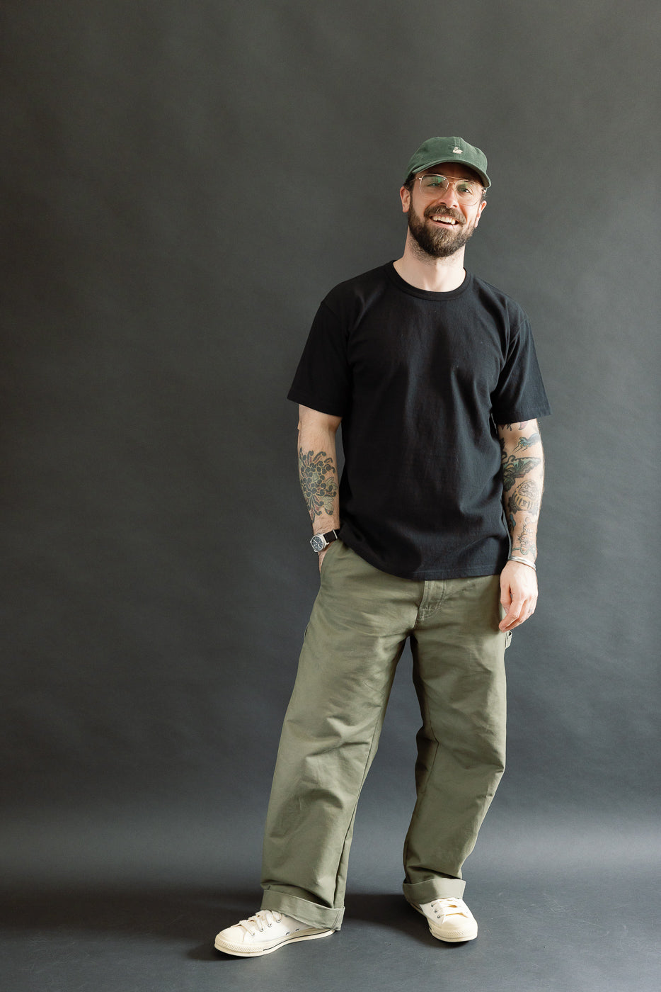 A Caucasian male model wearing the ELS Recycled Cotton Slub Crewneck T-Shirt - Black, showcasing its modern athletic fit with rolled-up sleeves, paired with loose olive-green trousers and off-white canvas sneakers.