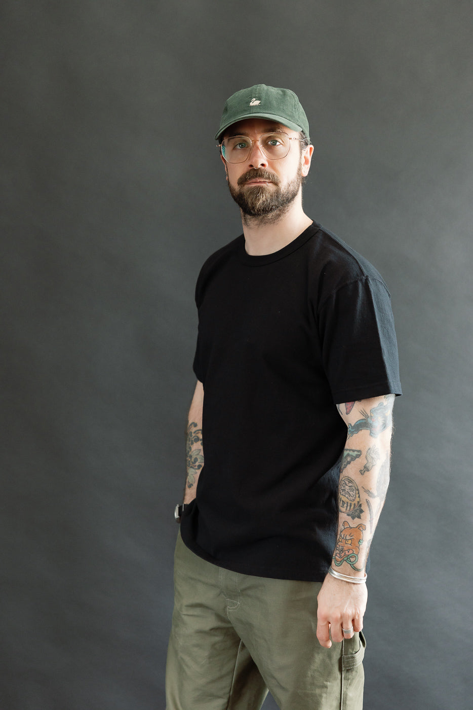 Bearded man in olive cargo pants and forest-green cap models the ELS Recycled Cotton Slub Crewneck T-Shirt - Black, showcasing its modern athletic fit and dense texture against a dark-gray backdrop.