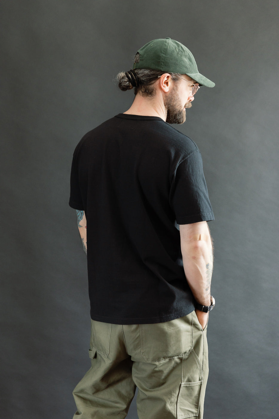 Man with back turned slightly right wears ELS Recycled Cotton Slub Crewneck T-Shirt - Black, showcasing relaxed fit and texture. Styled with olive cargo pants, embodying Dant's premium heritage-inspired menswear aesthetic.