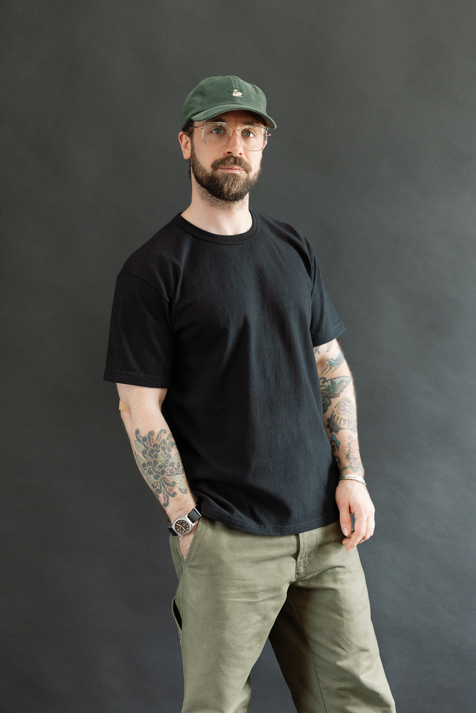 A man models the ELS Recycled Cotton Slub Crewneck T-Shirt - Black, showcasing its modern athletic fit and textured finish, with clear-framed glasses and a dark green baseball cap.