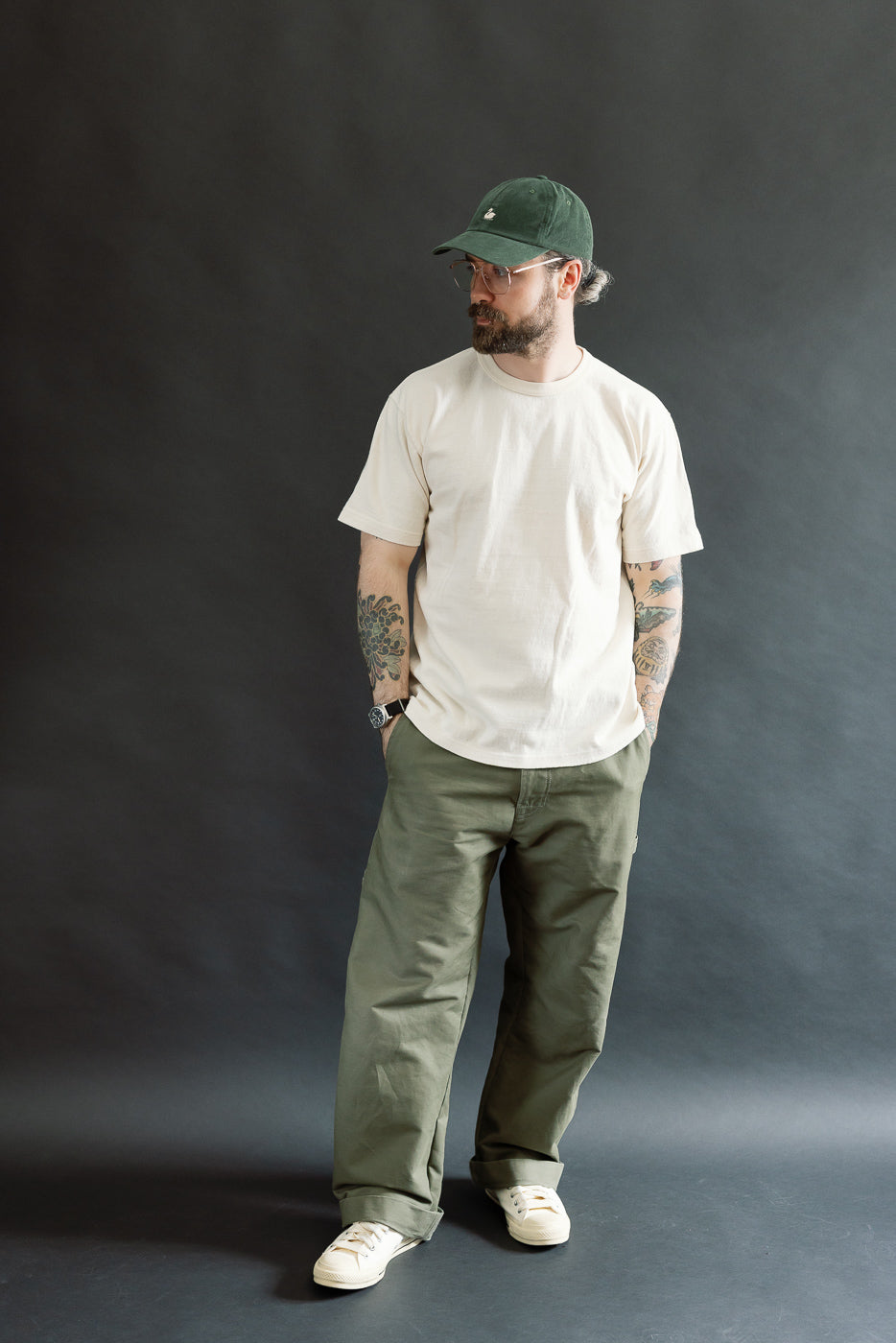 A Caucasian male model wearing the ELS Recycled Cotton Slub Crewneck T-Shirt - Ecru, styled with loose olive-green trousers and off-white sneakers, showcasing a casual, heritage-inspired look.
