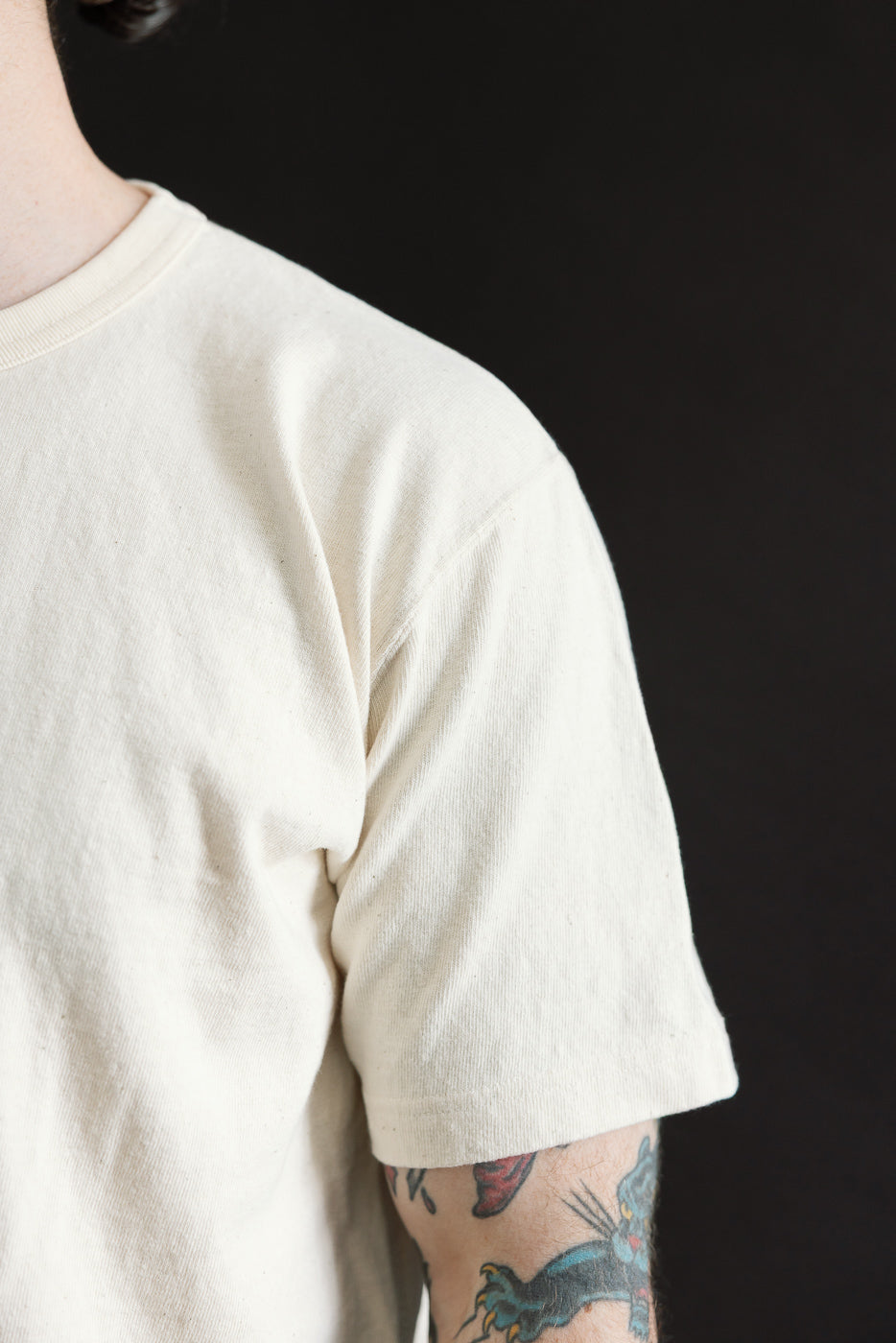 Close-up of a forearm tattoo peeking from under the ELS Recycled Cotton Slub Crewneck T-Shirt - Ecru, highlighting the shirt's textured, soft fabric and modern athletic fit against a black background.