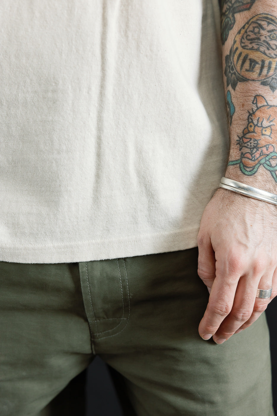 Close-up of the ELS Recycled Cotton Slub Crewneck T-Shirt - Ecru, showcasing its soft, textured fabric and modern athletic fit, worn over olive-green pants. Visible tattoos and accessories on the model's arm.