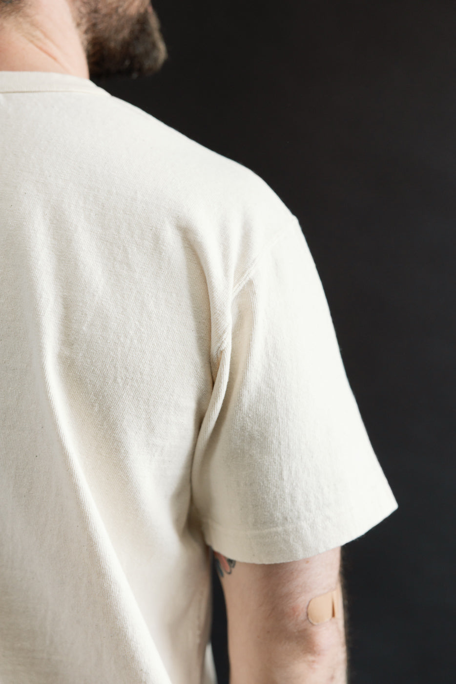 Person wearing ELS Recycled Cotton Slub Crewneck T-Shirt - Ecru, showcasing the upper back and right shoulder, with a visible bandage on the elbow against a dark background.