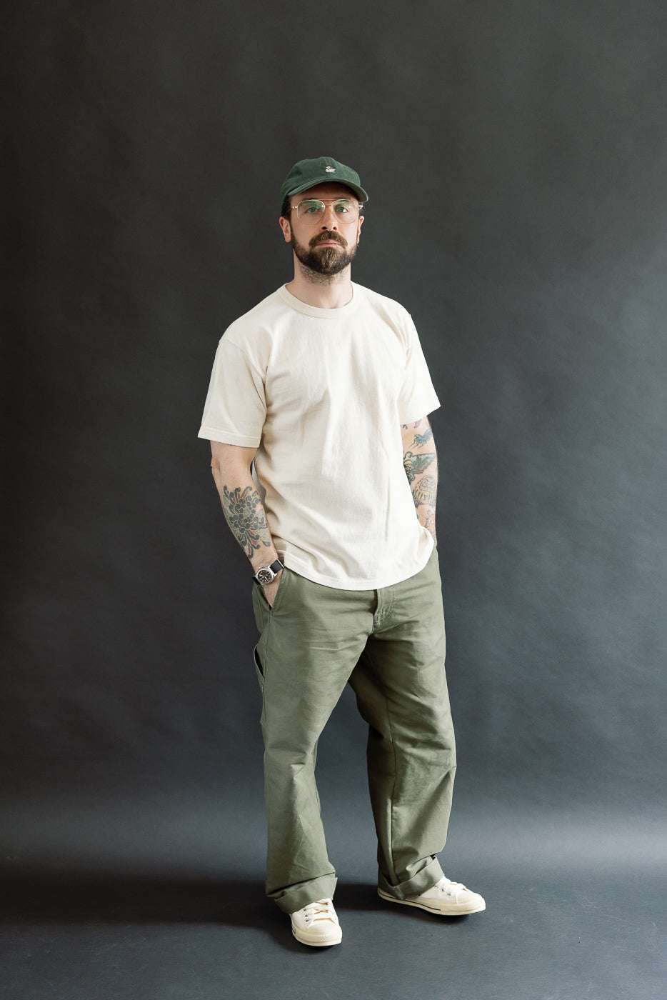 A full-length portrait of a man wearing the ELS Recycled Cotton Slub Crewneck T-Shirt - Ecru, showcasing its relaxed fit, tucked into olive-green pants, styled with clear-lens glasses and a baseball cap.