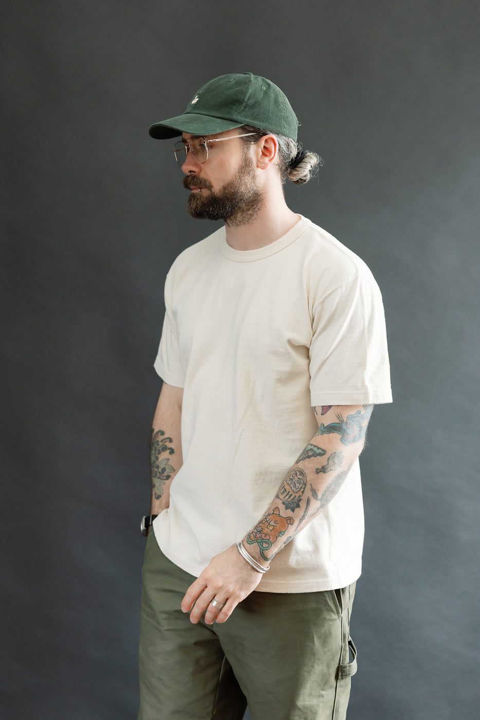 The image features a model wearing the ELS Recycled Cotton Slub Crewneck T-Shirt - Ecru, showcasing its athletic fit and textured fabric. He's styled with olive cargo pants and a dark green baseball cap.