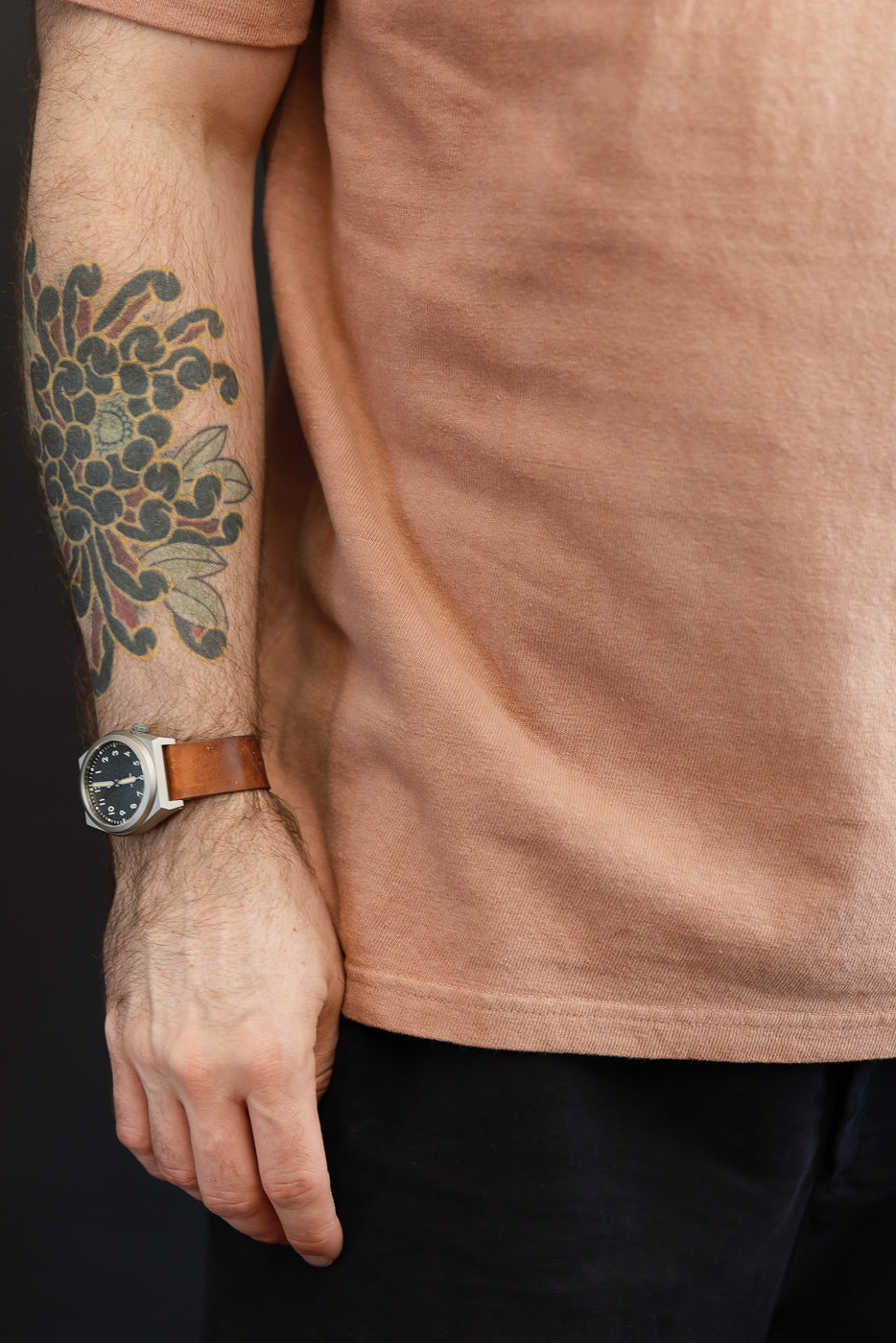 Close-up of male model's arm in tan tee, showcasing tattoo, watch, and fabric texture.