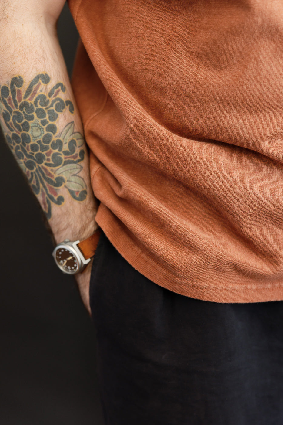 Close-up side view of male model's torso in rust tee, showing sleeve hem, fabric texture, tattooed arm, and watch.