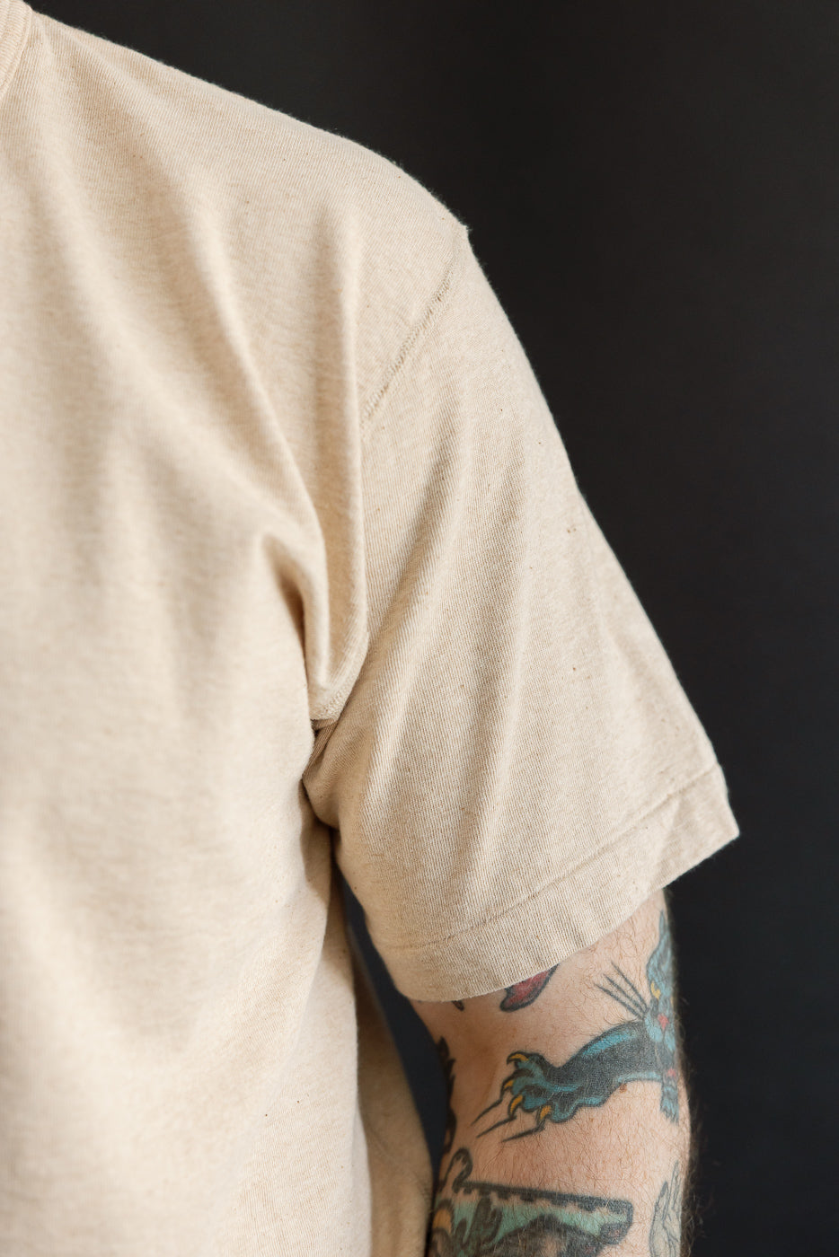 Close-up of male model’s left shoulder & sleeve, beige tee, ribbed hem, athletic build, tattooed arm