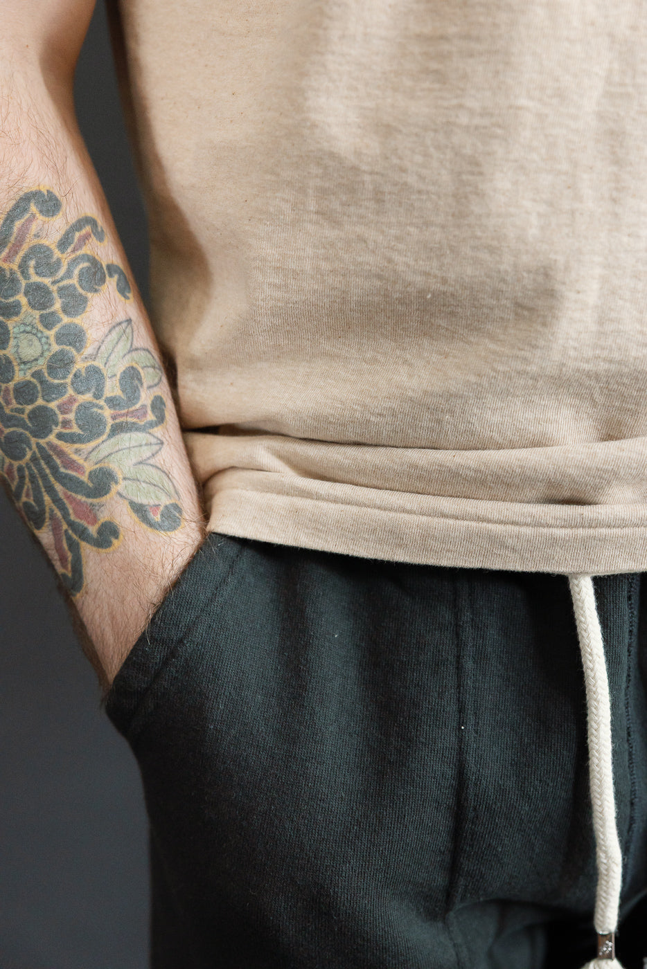 Close-up of male model's side, showing beige tee hem and dark drawstring pants, tattoo visible on arm