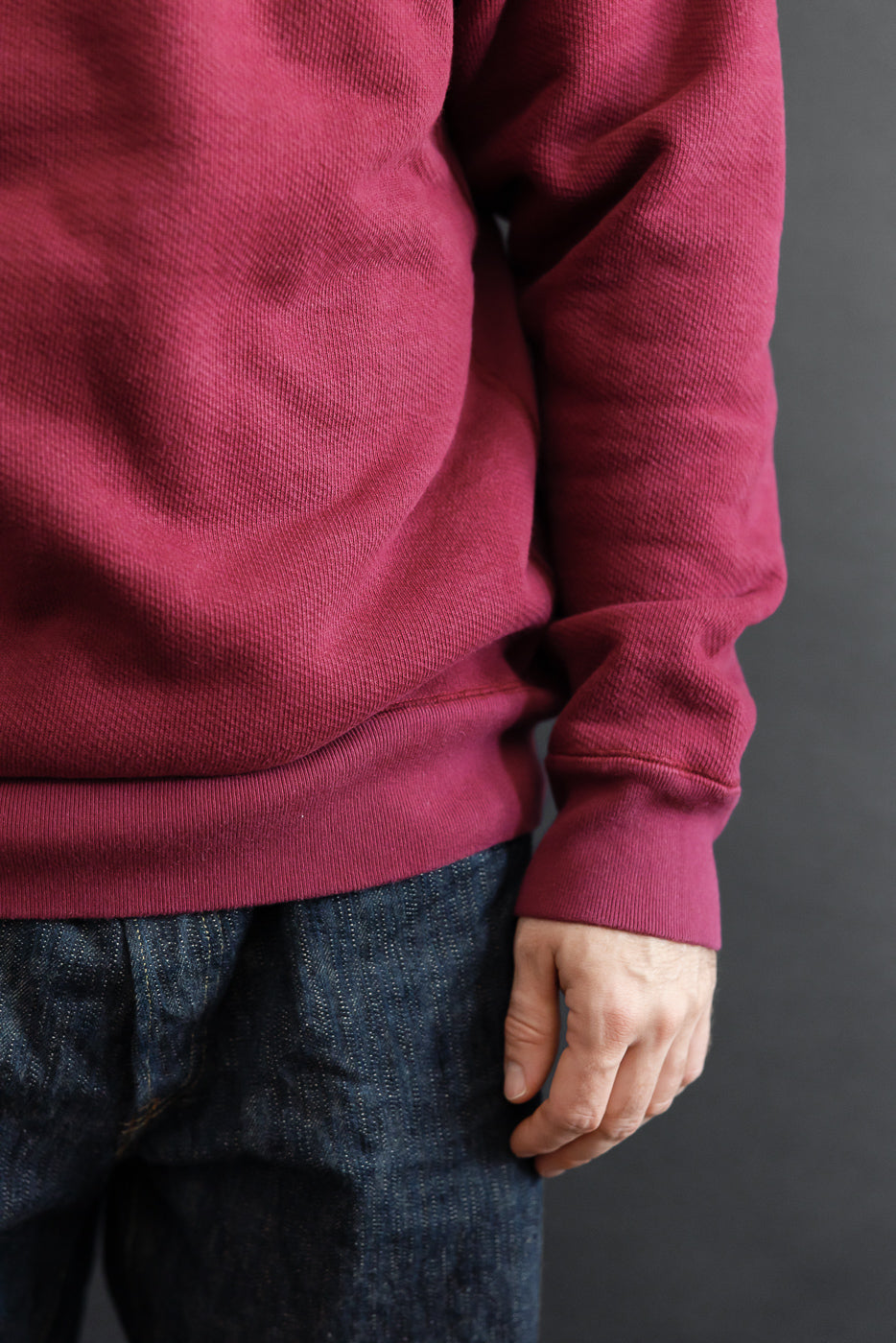 Close-up side view of a male model's burgundy waffle knit sweatshirt and dark selvedge denim.