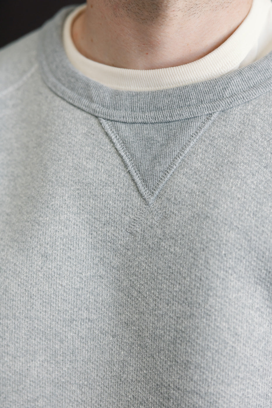 Close-up of the front crewneck collar and V-stitch on a grey sweatshirt worn by a male model.
