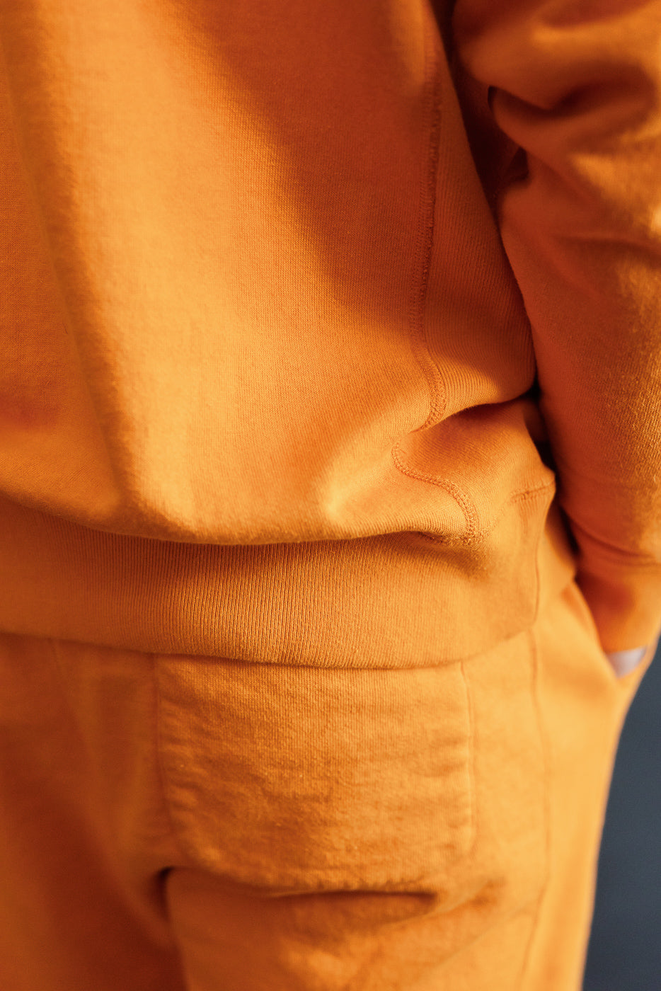 Close-up side view of male model's orange hoodie & joggers, showing ribbed hem, pocket detail, and soft fabric texture