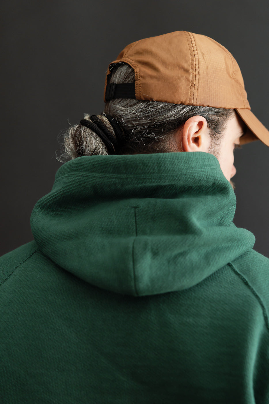 Close-up back view of a male model wearing a green cotton hoodie and tan cap.