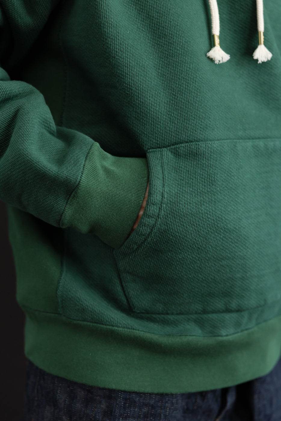 Close-up of the front pocket and ribbed cuff on a green cotton hoodie worn by a male model.