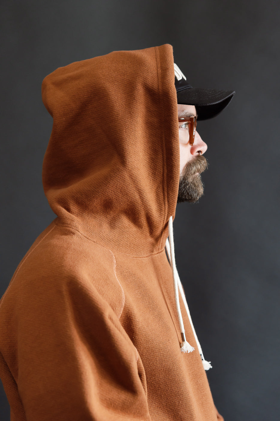 Side close-up of men's rust French terry pullover hoodie, hood up, on athletic 5'9