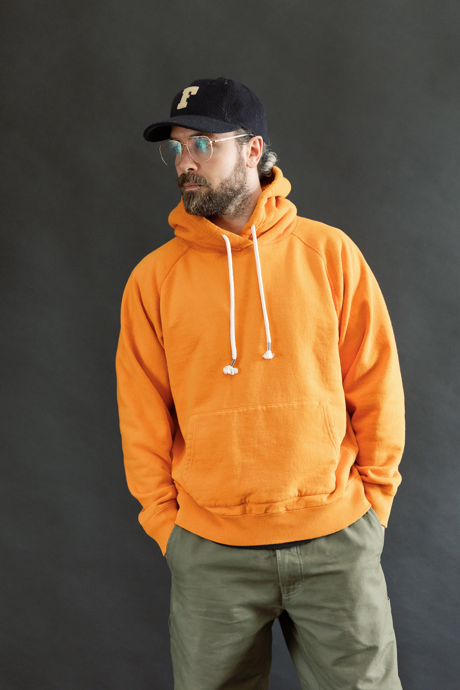 Bearded man models the Pullover Hoodie - Super Looper French Terry - Orange with white drawstrings, showcasing its modern athletic fit against a dark gray backdrop.