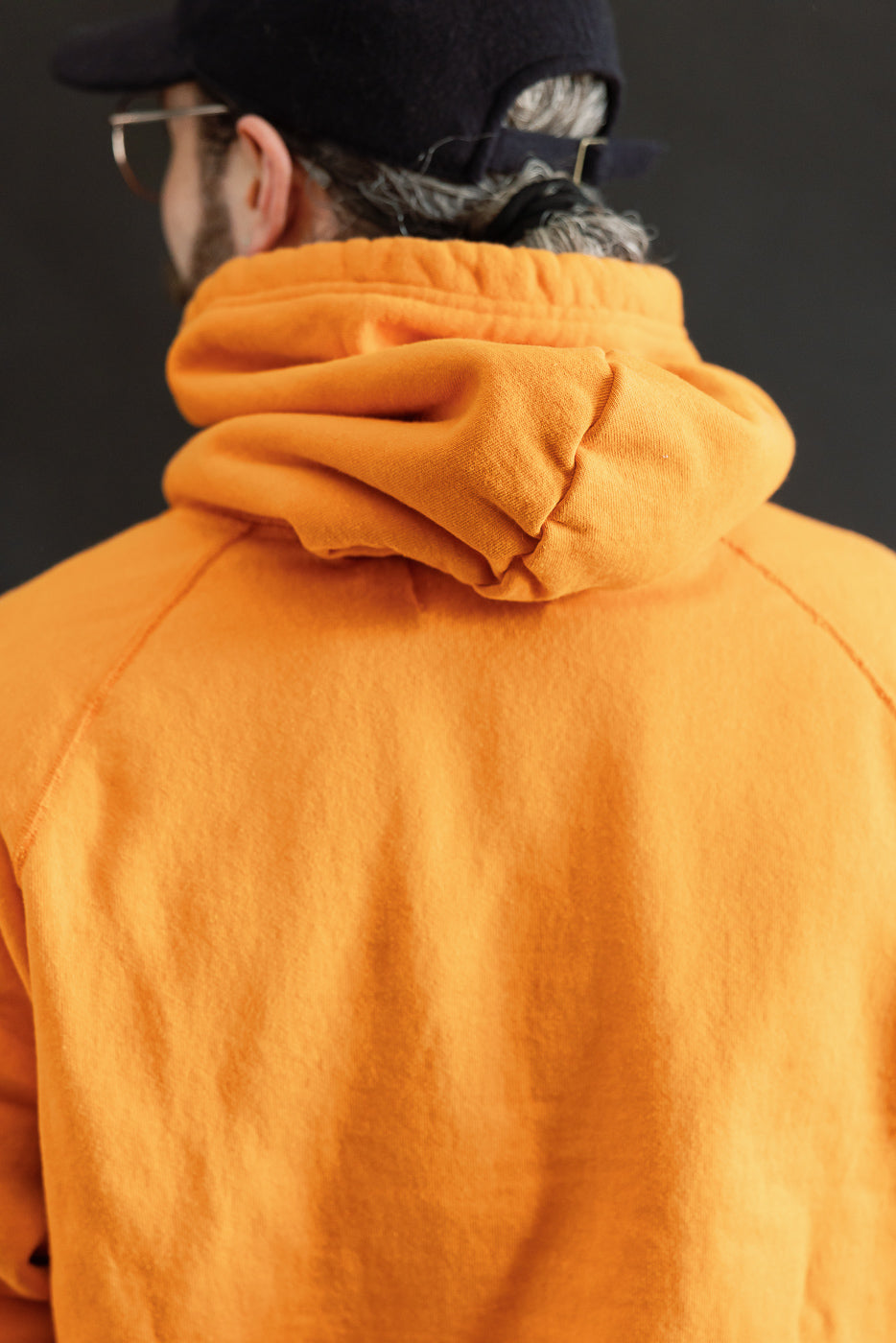 Person in bright orange Pullover Hoodie - Super Looper French Terry - Orange, hood down, black cap, gray hair visible, against dark background, showcasing innovative Japanese fabric texture and athletic silhouette.