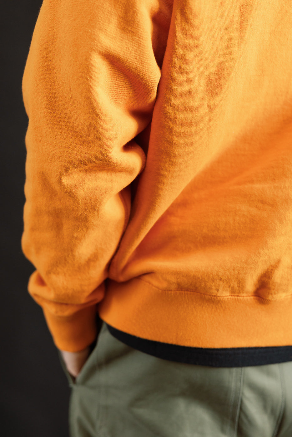 Close-up of the Pullover Hoodie - Super Looper French Terry - Orange, showcasing its smooth exterior and unique loopback texture, with hand in pocket of olive pants.