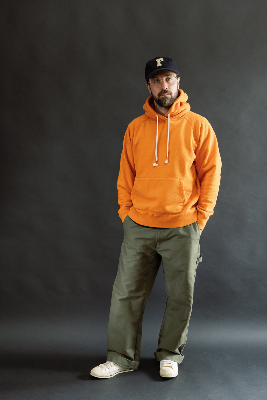 Man wearing Pullover Hoodie - Super Looper French Terry - Orange, featuring white drawstrings, paired with olive-green pants, off-white sneakers, and a dark baseball cap, posed against a dark-gray backdrop.