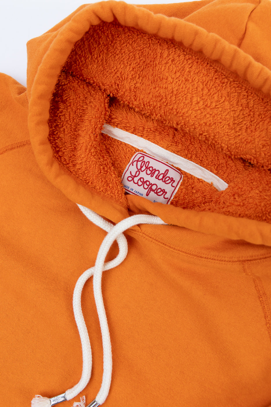 Close-up of the Pullover Hoodie - Super Looper French Terry - Orange, showcasing the terry-cloth lined hood, white drawcords, and Wonder Looper – Made in Japan woven label.