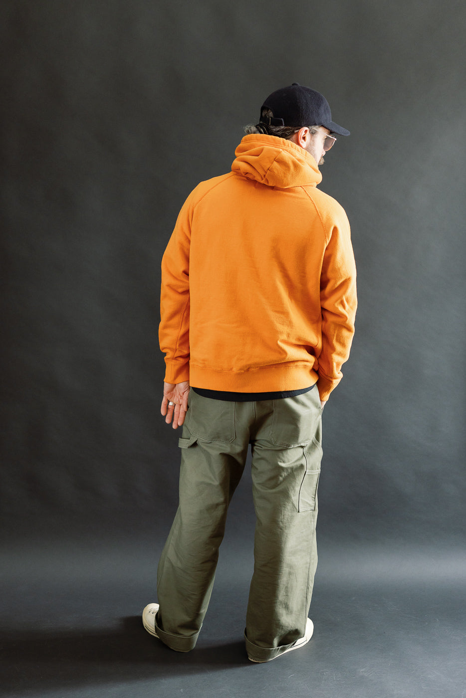 Model wearing Pullover Hoodie - Super Looper French Terry - Orange from behind, paired with olive cargo pants and white sneakers, showcasing its smooth exterior and athletic fit against a dark backdrop.