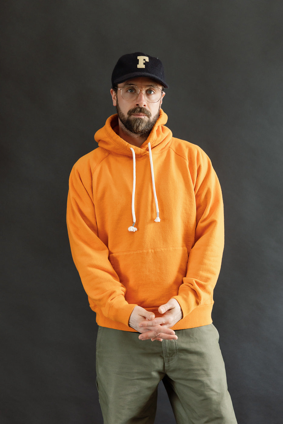 A bearded man models the Pullover Hoodie - Super Looper French Terry - Orange, showcasing its bright hue, drawstring hood, and kangaroo pocket. He pairs it with olive-green pants and a baseball cap.