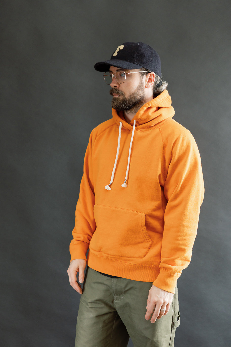 Man in a bright orange Pullover Hoodie - Super Looper French Terry, showcasing a modern athletic fit with white drawstrings and kangaroo pocket, paired with olive-green pants, against a plain backdrop.