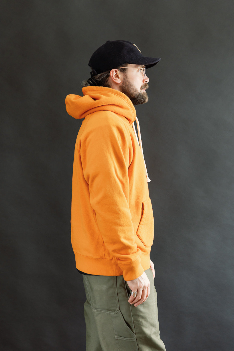 Man in profile wearing the Pullover Hoodie - Super Looper French Terry - Orange, showcasing its smooth exterior and modern athletic fit. Hood up, paired with olive cargo pants and black cap.